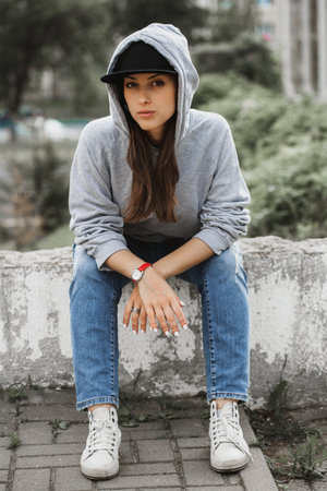 pretty serious brunette sitting, wearing grey baize, jeans, white gumshoes, black capの写真素材