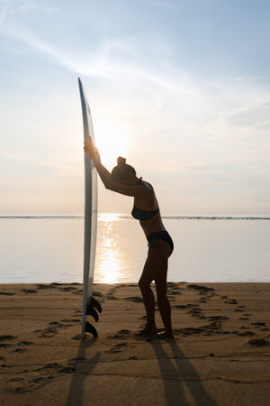 young pretty girl or fit sporty woman silhouette on sunny beach holding surfboard, looking down. water (ocean or sea) on backgroundの写真素材