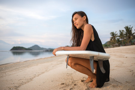 smiling happy excited active girl or woman holding surfboard on her knees, looking at water. awesome nature, warm weather, tropical atmosphereの写真素材