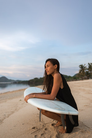 sun-tanned brunette attractive girl or woman (tourist, surfer) sitting with white surfboard on sandy beach near sea or ocean. holiday mood, warm weatherの写真素材