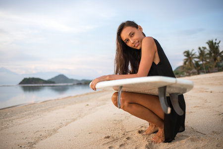 cute sporty surfer girl or smiling happy brunette woman with surfboard relaxing, having rest on sunny beach near the ocean or sea. mountains, palms on backgroundの写真素材