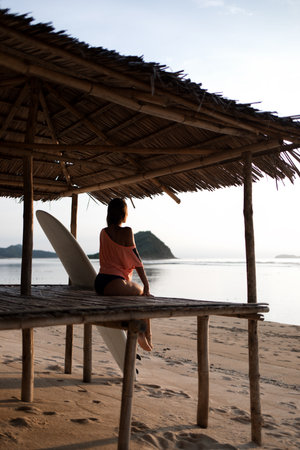 dreaming romantic sporty girl or woman (model, surfer) sitting on sandy sunny beach near ocean or sea, looking at water, restingの写真素材