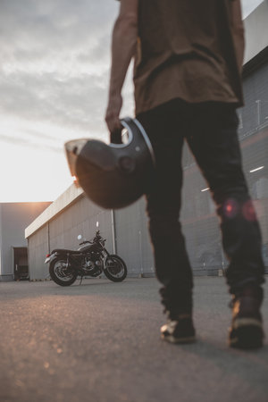 Close-up of a handsome rider biker guy hand with black helmet in front of classic style cafe racer motorcycle. Bike custom made in vintage garage. Brutal fun urban lifestyle. Outdoor portrait.の写真素材