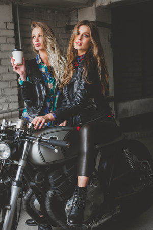 Two sexy biker girls sitting on cafe racer motorcycles. Attractive women with healthy long hair wearing stylish leather jackets sit on bikesの写真素材