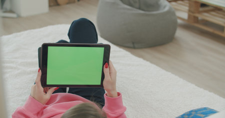 View over head on of caucasian woman lying on the bed at home. Girl holding tablet computer with green screen while watching something. Chroma key.の写真素材