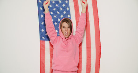 Pretty stylish girl in pink hoodie celebrating independence day and having fun over national usa flag on white wall on background.の写真素材