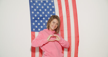 Pretty stylish girl in pink hoodie celebrating independence day and having fun over national usa flag on white wall on background. Young woman show heart sign symbolの写真素材