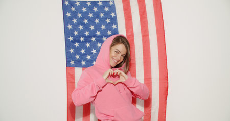 Pretty stylish girl in pink hoodie celebrating independence day and having fun over national usa flag on white wall on background. Young woman show heart sign symbolの写真素材