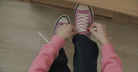 Young woman skateboarder tying shoelace in pink sneakers. Close-up of legs of girl sitting on chair at home and getting ready for walking. Top view の写真素材