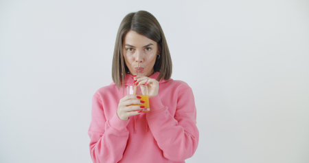 Caucasian young girl in sunglasses and stylish outfit drinking juice with glass straw and smiling over white wall background.の写真素材