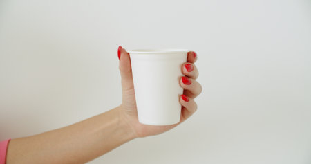 Mockup of female hand holding coffee paper cup on light white wall background