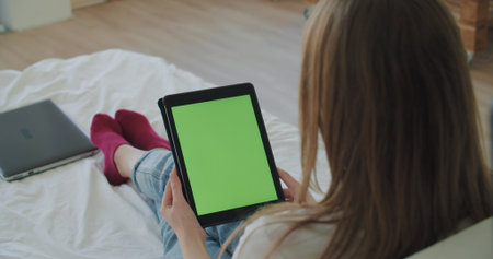 View over head on of caucasian woman lying on the bed at home. Girl holding tablet computer with green screen while watching something. Chroma keyの写真素材
