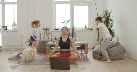 Mother meditating with daughter while active energetic child son playing, Mom working and doing yoga exercise at home for stress relief relaxing with naughty little kid.の写真素材