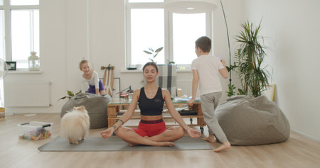 Mother meditating with daughter while active energetic child son playing, calm young mom doing yoga exercise at home for stress relief relaxing with naughty little kid.の写真素材