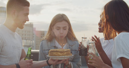 Young woman birthday girl is making wish, blowing candle on cake and clapping hands while her friends are congratulating her during rooftop partyの写真素材
