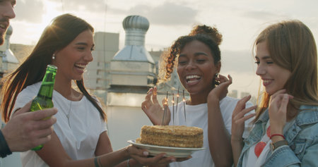 Young woman birthday girl is making wish, blowing candle on cake and clapping hands while her friends are congratulating her during rooftop partyの写真素材