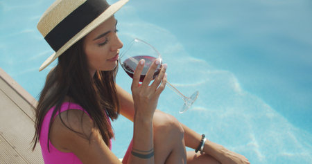 Smiling woman sitting on the pool edge with glass of wine. Beautiful woman relaxing outdoors at summer sunny dayの写真素材