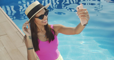 Young woman in hat and sunglasses, making selfie, showing tongue, funny face, posing at pool at sunny summer day, stylish hatの写真素材