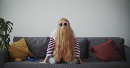 Funny young woman having fun on sofa in morning. Attractive red girl in cheerful mood . Female model in home clothes waving her hair and wearing sunglasses.の写真素材