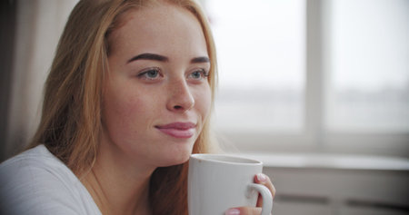 Close-up of face of young caucasian woman smiling and drinking hot cocoa or coffee in morning light....