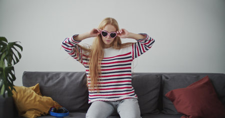 Funny young woman having fun on sofa in morning. Attractive red girl in cheerful mood . Female model in home clothes waving her hair and wearing sunglassesの写真素材