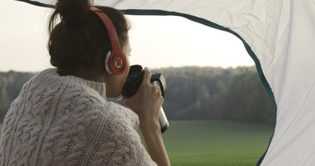 Camping woman lying in tent . Close up of girl relaxing on vacation. Young happy female caucasian model in headphones listening to music and drink coffeeの写真素材