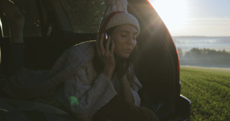 Happy girl relaxing on vacation. Young female caucasian model in headphones listening to music and reading texting message on smart phone in trunk of car. Woman drinking hot coffee or teaの写真素材