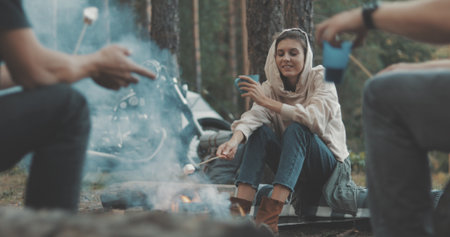 Group of happy friends around burning camping bonfire in woods roasting marshmallows talking and smiling. Hikers sitting around camp fire. Outdoor picnic in forest.の写真素材