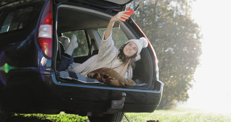 Young woman admiring the sunrise in trunk of car. Girl dressed taking selfie photo on mobile phone against backdrop of forest panorama in fogの写真素材