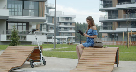Woman sitting on bench among urban space and reading ebook working using digital tablet. Attractive girl with electric kick scooterの写真素材