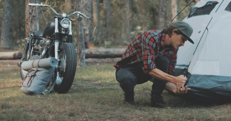Hiker camping in forest. Guy biker sets up tent in woods. Handsome young man tourist puts hiking backpack. Countryside lifestyle scene.の写真素材