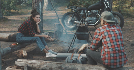 Hiker friends sitting around camp fire. Outdoor camping picnic in forest. Freedom lifestyle scene. Young couple in love sit by fireplace and talkの写真素材