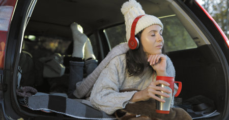 Happy girl relaxing on vacation. Young female caucasian model in headphones listening to music and reading texting message on smart phone in trunk of car. Woman drinking hot coffee or teaの写真素材