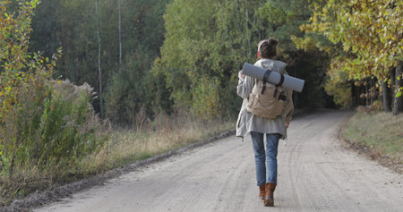 Active healthy female hipster model hiking along forest mountain road. Caucasian girl rest outdoors . Woman tourist walking in fall park at sunny autumn day. Back viewの写真素材
