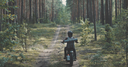 Young rider driving vintage classic custom motorcycle on forest road. Tourist with backpack on back goes to rest in campの写真素材