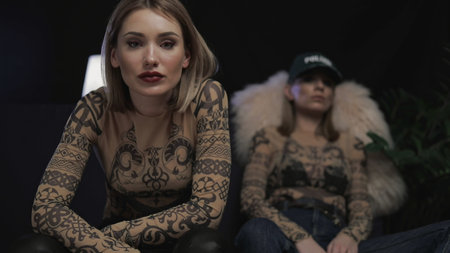 Two fashion young women sit on sofe and look at camera at dark room. Indoors lifestyle