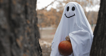 Little girl in a costume ghost with orange pumpkin in autumn park or forest. Halloween celebration holiday. Funny smiling grimaceの写真素材