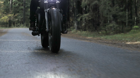 A guy in a black leather jacket and helmet riding a classic motorcycle on a forest road.