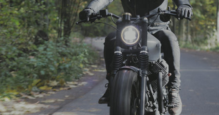 A guy in a black leather jacket and helmet riding a classic motorcycle on a forest road.