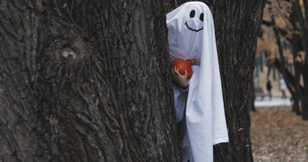 Little girl in a costume ghost with orange pumpkin in autumn park or forest. Halloween celebration holiday. Funny smiling grimaceの写真素材