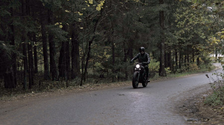 A guy in a black leather jacket and helmet riding a classic motorcycle on a forest road.