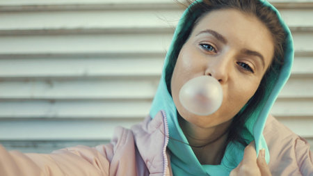 Outdoor lifestyle portrait of beautiful young woman blowing a bubble gum balloon. Urban city lifestyleの写真素材