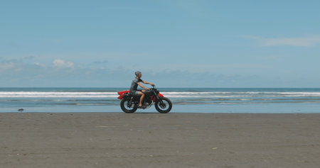 Handsome man biker surfer driving his black motorbike cafe racer with red surfboard shortboard on the beach along the ocean at sunny dayの写真素材