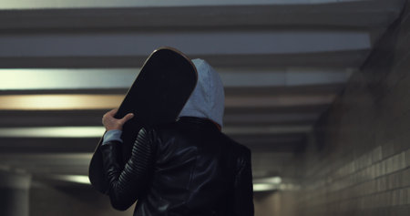 Girl teenager goes at night in the underpass. Young caucasian woman with skateboard in dark underground tunnel. Urban city lifestyle. Back viewの写真素材