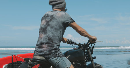 Handsome man biker surfer driving his black motorbike cafe racer with red surfboard shortboard on...