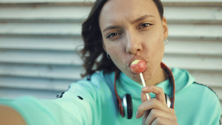 Pretty smiling girl licking lollipop and making selfie using smartfone. Close-up portrait young beautiful caucasian woman holding phone taking self photoの写真素材
