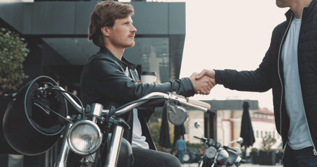 Two friends motorcyclists men meet on the street and shake hands with each other. Guys bikers in...