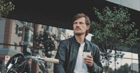 Young caucasian man biker drinking coffee while standing near the classic vintage motorcycle from...