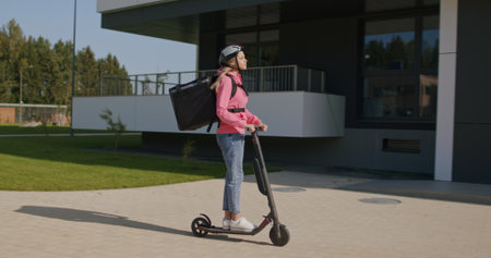 Food delivery female courier wearing hoodie rides electric scooter to deliver orders and packages...