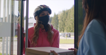 Female customer opens door to pizza deliverman. Courier wearing in protective medical mask. Express...
